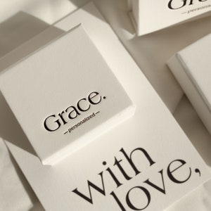 May include: A square off-white gift box with the black text Grace. -personalized- printed on the lid. Below the box, a card features the black text with love, in a serif font. The box has a textured surface and is positioned at an angle.