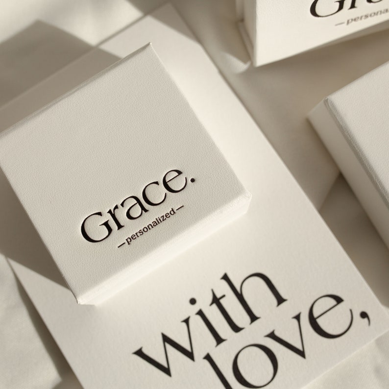 May include: A square off-white gift box with the black text Grace. -personalized- printed on the lid. Below the box, a card features the black text with love, in a serif font. The box has a textured surface and is positioned at an angle.