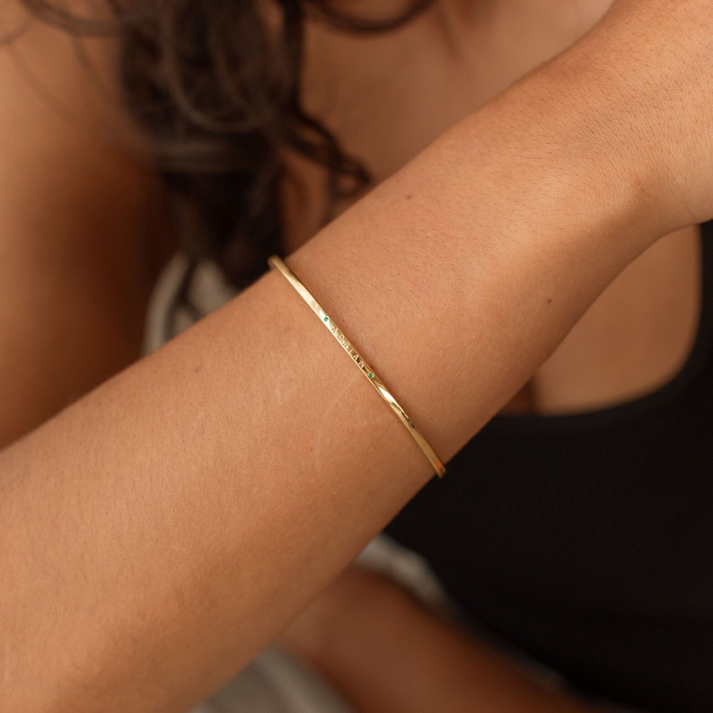 A gold bangle bracelet with a small emerald green gemstones embedded along its length. The minimalist design is simple and elegant, suitable for everyday wear. The bracelet is shown on a wrist, highlighting its delicate and comfortable fit.