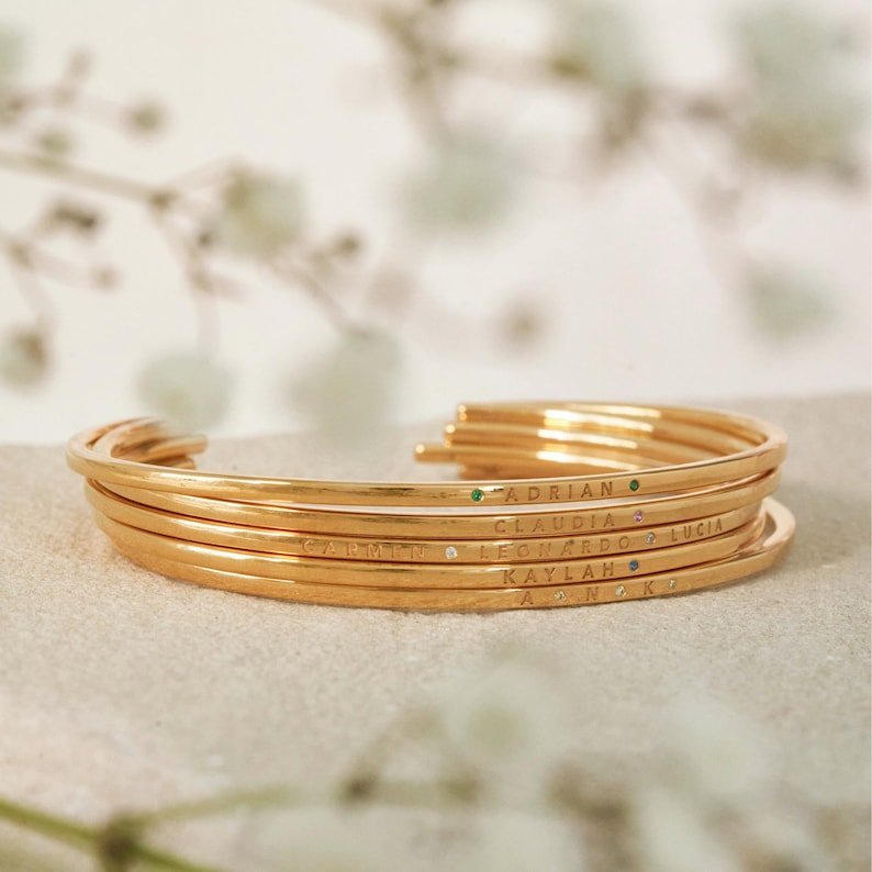 Close-up of gold stacked bangles with engraved names: Adrian, Claudia, Lucia, Leonardo, Kaylah, and Anok. Each name is separated by a small gemstone; emerald green, pink, and clear. The bangles are thin and delicate, ideal for layering.