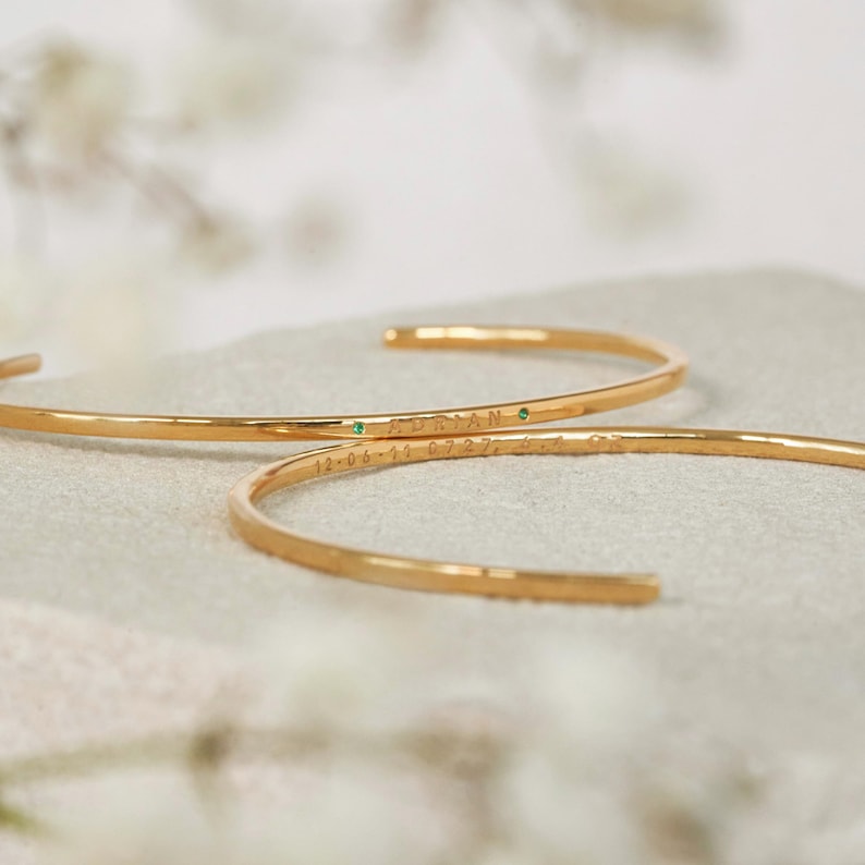 Close-up of two gold bangle bracelets. Each bracelet features engraved text including names and dates, and two small emerald green gemstones. The bracelets are delicate and minimalist in style.