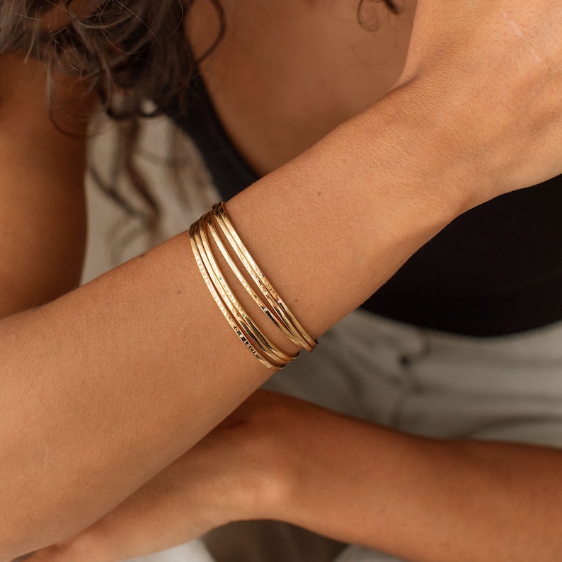 Close-up of several gold bangles stacked on a wrist.  They appear to be made of metal with a high polish finish. The gold tone is consistent across all bangles. The design is simple and elegant, suitable for layering.