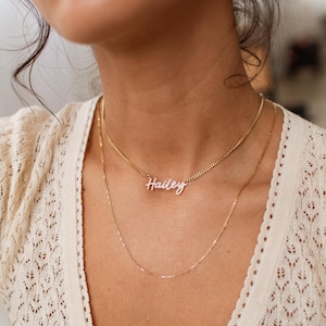 May include: A gold-colored necklace with the name "Hailey" in white script lettering. The necklace is layered with a delicate chain. The model is wearing a cream-colored, open-knit sweater.