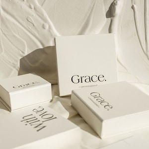 Op de afbeelding: Vier witte dozen met het woord "Grace" erop gedrukt. De dozen zijn op elkaar gestapeld en staan op een wit oppervlak. De dozen zijn voor gepersonaliseerde geschenken.