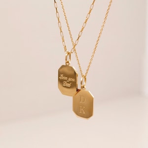 Two gold necklaces with octagonal pendants. One pendant is engraved with 'Love you, Dad.' The other pendant is engraved with the initials 'DK'.  Both necklaces feature delicate gold chains.