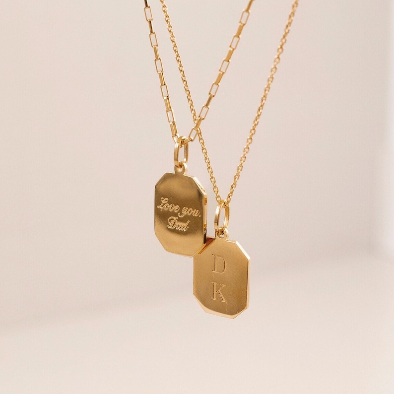 Two gold necklaces with octagonal pendants. One pendant is engraved with 'Love you, Dad.' The other pendant is engraved with the initials 'DK'.  Both necklaces feature delicate gold chains.