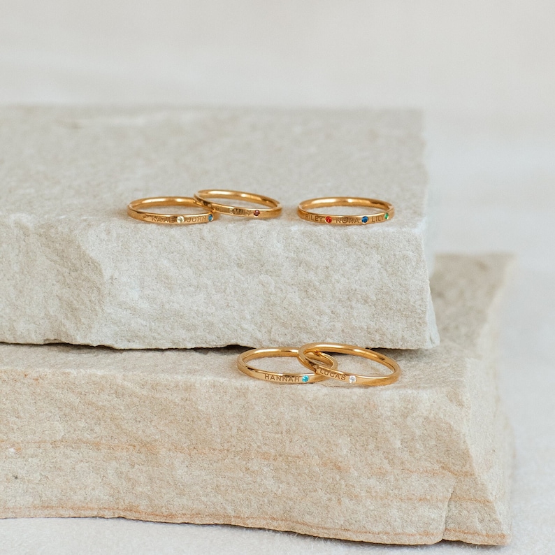 Stack of five gold name rings with small gemstones, engraved with names and placed on a stone background
