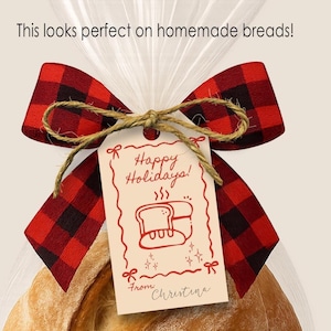 May include: A loaf of golden-brown bread in a clear cellophane bag, tied with a red and black plaid bow and twine. A small tag reads "Happy Holidays!" and "From Christina."