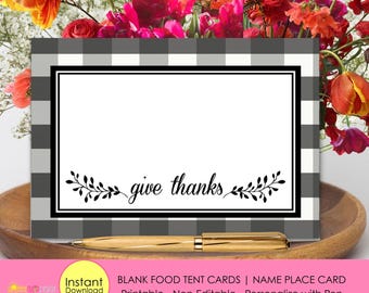 May include: A rectangular card featuring a gray and white buffalo check border, a central white space, and the black text "give thanks" flanked by decorative leaf illustrations. A gold-toned pen rests horizontally in front of the card.
