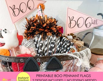 May include: Two triangular light pink pennant flags featuring the text BOO and BOO y'all in black script. The flags are displayed in a metal basket filled with Halloween-themed items including black and white striped straws and a ghost figure.