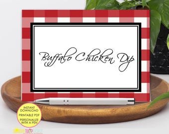 May include: A rectangular food label card featuring a red and white buffalo check border and a white center panel with the black text Buffalo Chicken Dip in a cursive script. A silver and white pen rests horizontally across the bottom edge.