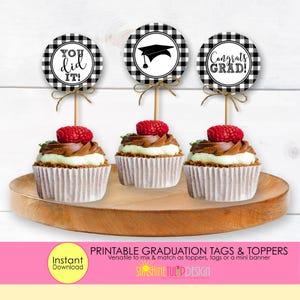 May include: Three cupcakes with graduation-themed toppers. Each cupcake has a raspberry on top of chocolate frosting and white frosting. The toppers are black and white checkered circles with graduation messages and a mortarboard cap. The cupcakes are on a wooden tray.