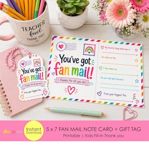 Op de afbeelding: Een witte invulbare fanmail-notitiekaart en cadeaulabel met een regenboogstempelontwerp. De kaart en het label hebben de tekst "You've got fan mail!" en "Thanks for all you do!". Een roze spiraalgebonden notitieboek, een mok met potloden en bloemen staan op de achtergrond.