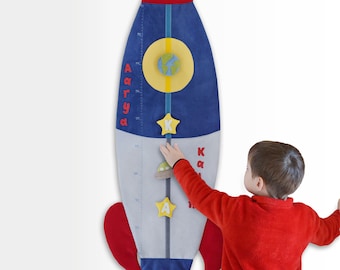 May include: A blue and red rocket-shaped growth chart with a yellow star, a green and blue planet, and a gray spaceship. The chart is marked with inches and has the name "Arya" written on it. A child is standing next to the chart and is touching the 110-inch mark.