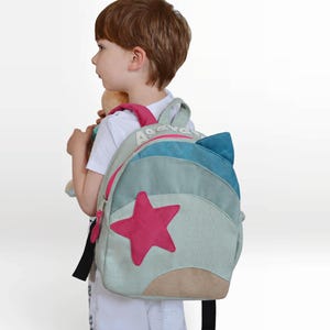 Peut inclure: Un sac à dos pour enfant avec un motif d'étoile. Le sac à dos présente une palette de couleurs bleu clair, sarcelle et rose. Une grande étoile rose est sur le devant. Le sac à dos a un design d'oreille de chat sur le dessus. Le mot "AS" est imprimé sur le dessus.