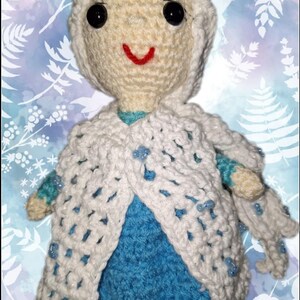 May include: A handmade crochet doll with a white hooded cape and a blue dress. The doll has black button eyes and a red smile. The cape is adorned with small blue beads. The background is a light blue and white floral pattern.