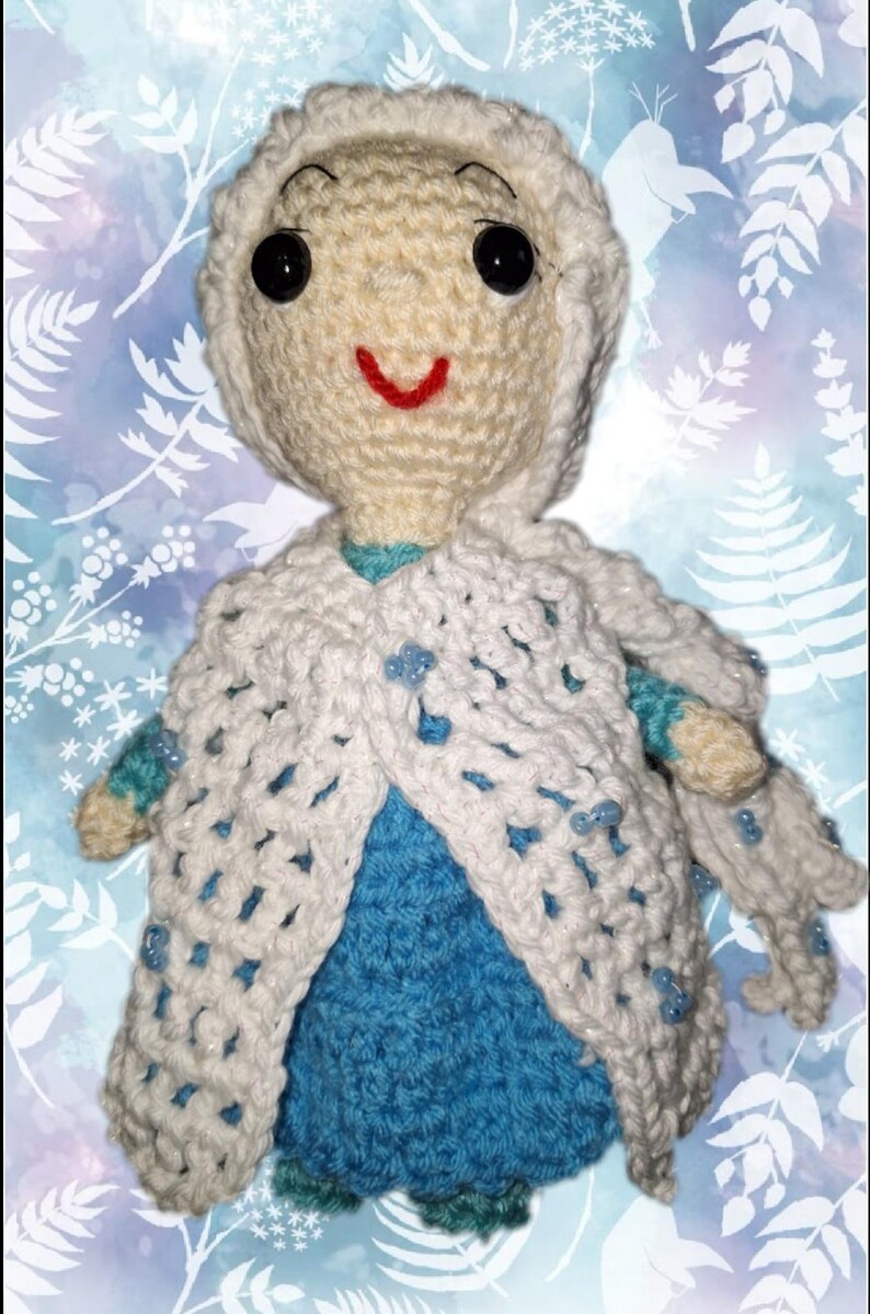 May include: A handmade crochet doll with a white hooded cape and a blue dress. The doll has black button eyes and a red smile. The cape is adorned with small blue beads. The background is a light blue and white floral pattern.