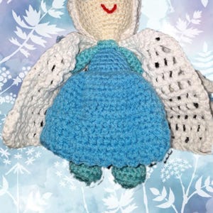 May include: A handmade crochet doll with a cream-colored head and a blue dress. The doll has black eyes and a red smile. It has white wings and teal shoes. The background is a blue and white floral pattern.