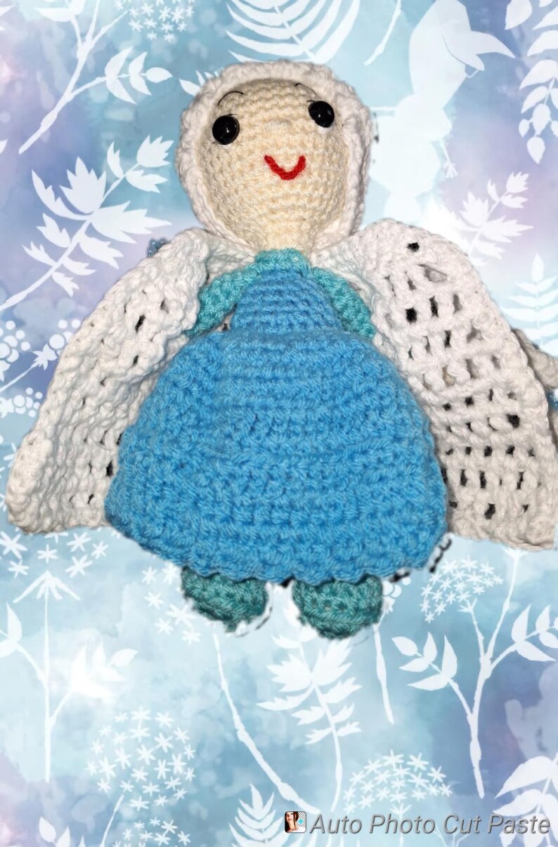 May include: A handmade crochet doll with a cream-colored head and a blue dress. The doll has black eyes and a red smile. It has white wings and teal shoes. The background is a blue and white floral pattern.