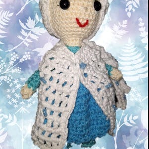 May include: A handmade crochet doll in the likeness of a character from a popular animated film. The doll features a cream-colored head and a white crocheted cape over a blue dress. The doll has black eyes and a red smile.