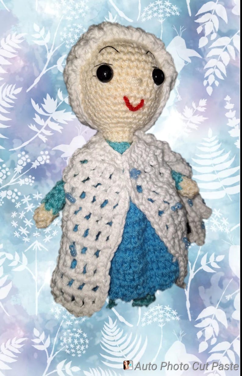 May include: A handmade crochet doll in the likeness of a character from a popular animated film. The doll features a cream-colored head and a white crocheted cape over a blue dress. The doll has black eyes and a red smile.