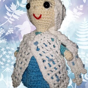 May include: A handmade crochet doll in the likeness of a princess, featuring a cream-colored face and hair, black eyes, and a red smile. The doll wears a blue dress and a white, open-weave cape adorned with blue beads. The background is a light blue with white floral patterns.