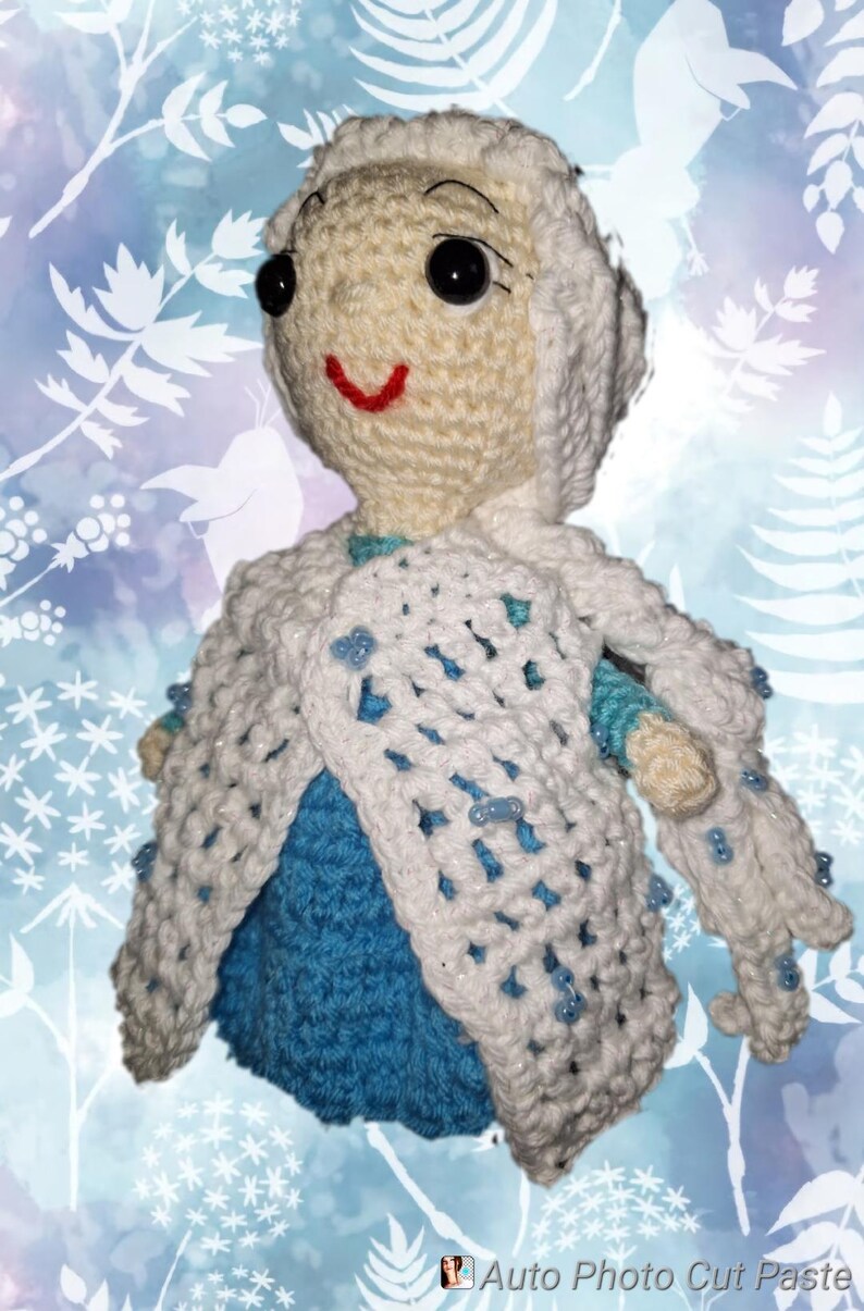 May include: A handmade crochet doll in the likeness of a princess, featuring a cream-colored face and hair, black eyes, and a red smile. The doll wears a blue dress and a white, open-weave cape adorned with blue beads. The background is a light blue with white floral patterns.