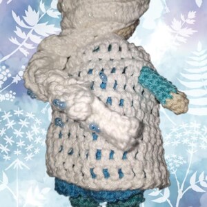 May include: A handmade crochet doll wearing a white hooded coat with blue accents. The coat has a textured, open weave design and is adorned with small blue beads. The doll's lower half is dressed in blue and green crochet.