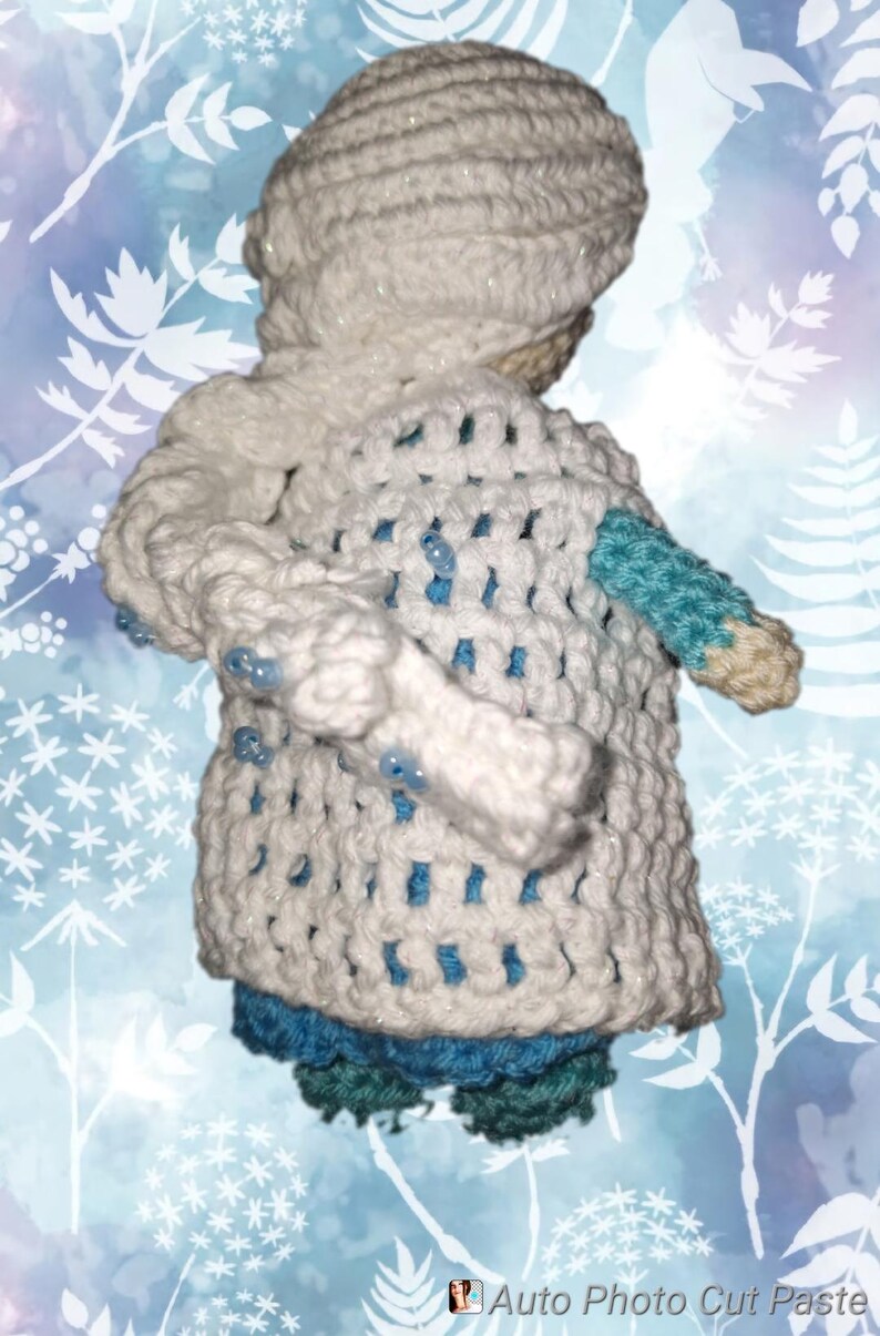May include: A handmade crochet doll wearing a white hooded coat with blue accents. The coat has a textured, open weave design and is adorned with small blue beads. The doll's lower half is dressed in blue and green crochet.
