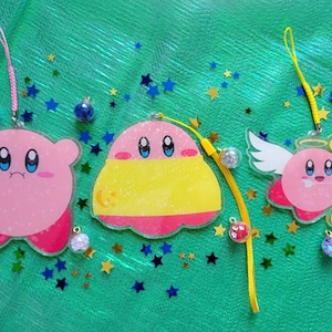 May include: Three pink and yellow character charms on clear strings. The charms are of a pink character with a sad expression, a pink character with a yellow dress and a pink character with wings and a bow and arrow.