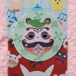May include: A collection of colorful mouse pads featuring cartoon characters and designs. The pads include a round green pad with a face design, a red pad with white and black characters, and a blue pad with musical notes and cartoon figures.
