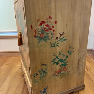 May include: Wooden cabinet with floral artwork in red, green, and blue. The cabinet has a light brown finish and a rectangular shape. A gold tassel hangs from the side. The cabinet is likely used for storage.