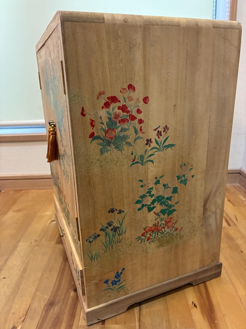 May include: Wooden cabinet with floral artwork in red, green, and blue. The cabinet has a light brown finish and a rectangular shape. A gold tassel hangs from the side. The cabinet is likely used for storage.
