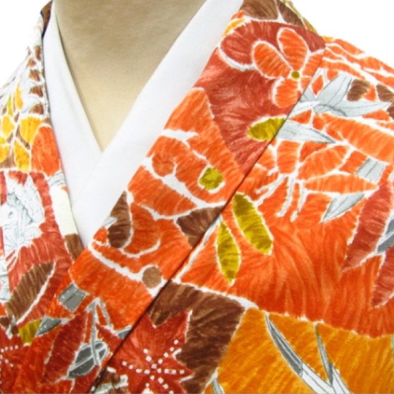 Puede incluir: Un primer plano de un kimono blanco con un estampado floral colorido. El estampado presenta flores y hojas de color naranja, marr&oacute;n y amarillo. El kimono est&aacute; doblado por la mitad, mostrando el cuello y la parte delantera de la prenda.