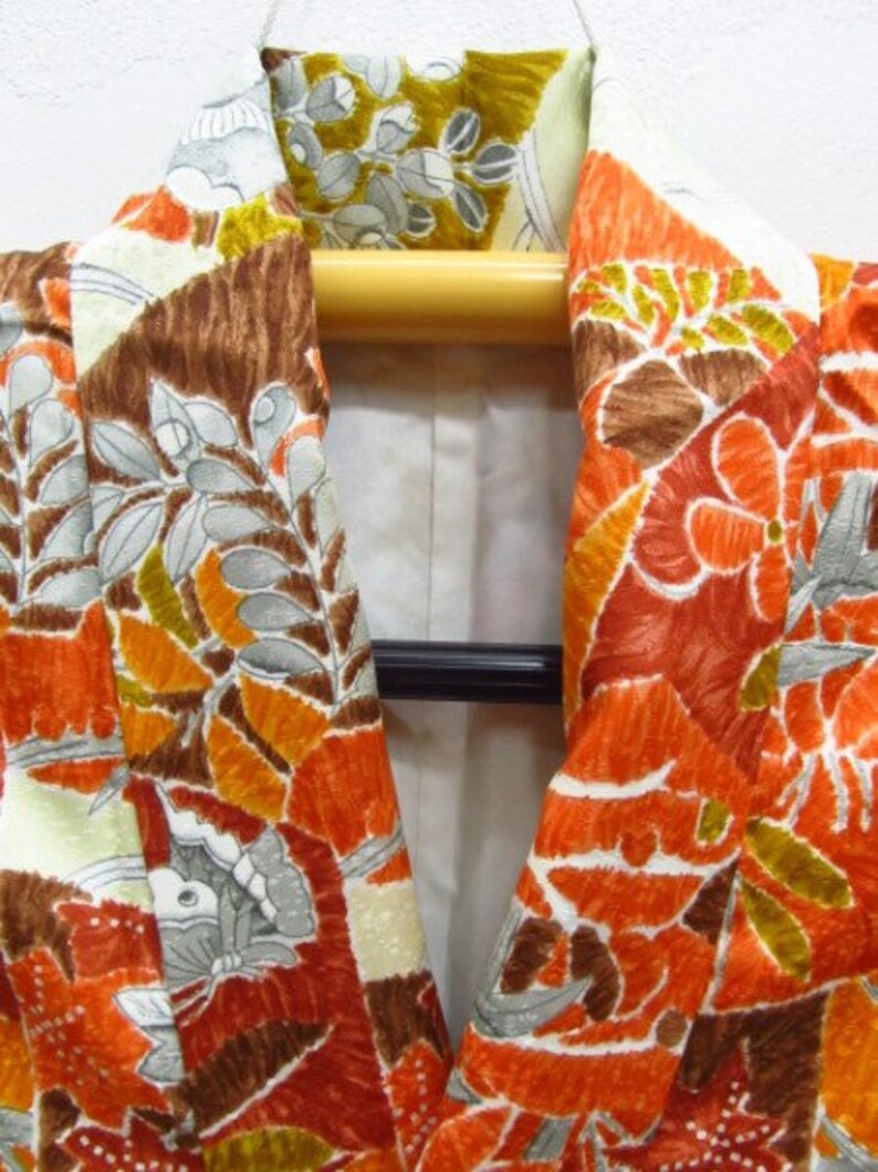 Puede incluir: Primer plano del cuello de un kimono marr&oacute;n, naranja y blanco con un patr&oacute;n floral. El kimono est&aacute; en una percha de madera.