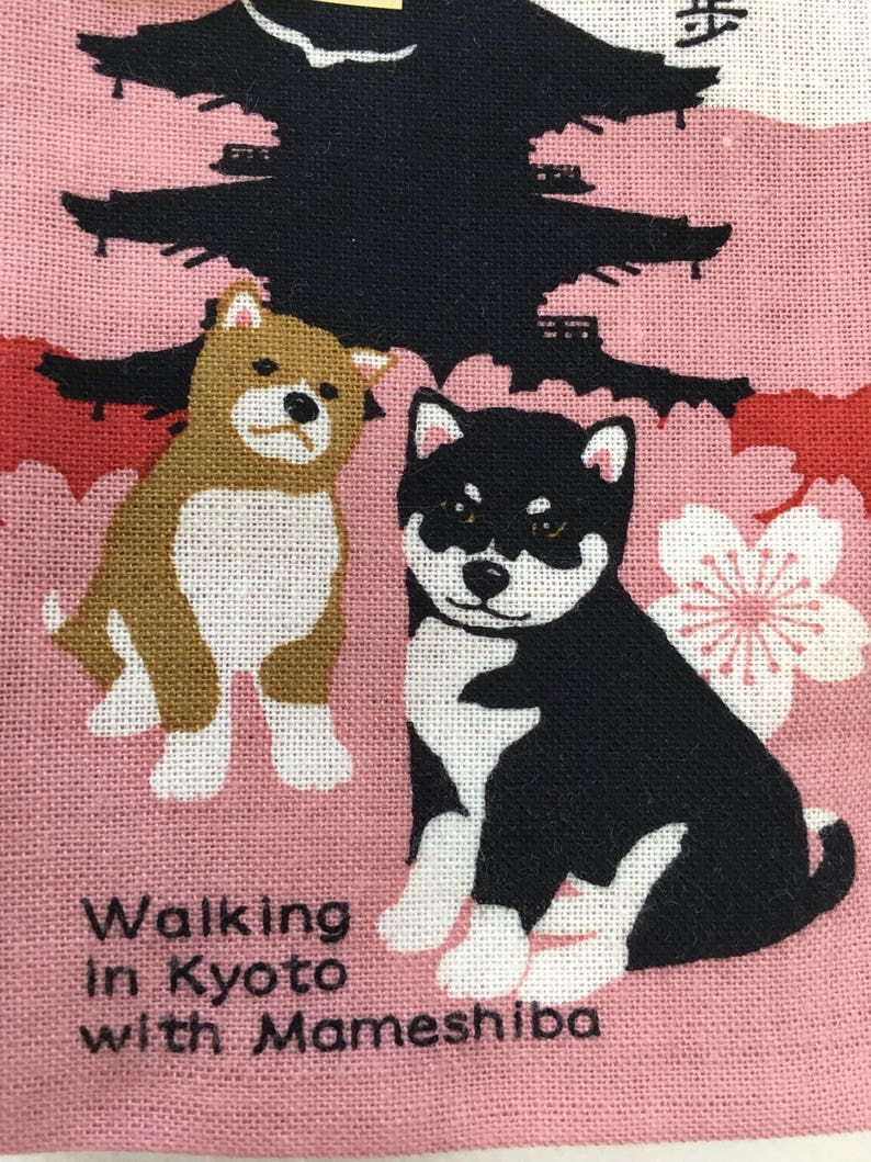 Op de afbeelding: Een roze stoffen tas met een zwart-witte illustratie van twee Shiba Inu honden. De honden lopen voor een zwarte silhouet van een pagode met roze kersenbloesems. De tekst "Walking in Kyoto with Mameshiba" staat op de tas gedrukt.