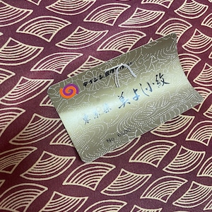 May include: A close-up of a burgundy fabric with a repeating pattern of gold-colored crescent shapes. A gold tag with Japanese text and the number "6500" is attached to the fabric.