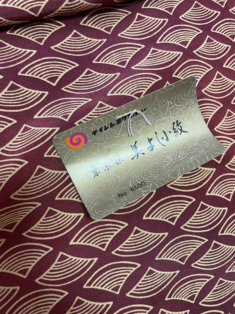 May include: A close-up of a burgundy fabric with a repeating pattern of gold-colored crescent shapes. A gold tag with Japanese text and the number "6500" is attached to the fabric.