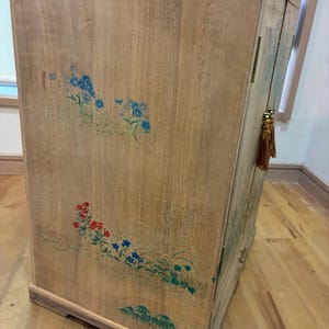 May include: Wooden cabinet with floral designs in blue, red, and green. The cabinet has a light brown finish and a curved top. A decorative tassel hangs from the side. The cabinet is a storage unit.