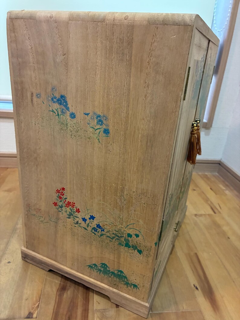 May include: Wooden cabinet with floral designs in blue, red, and green. The cabinet has a light brown finish and a curved top. A decorative tassel hangs from the side. The cabinet is a storage unit.
