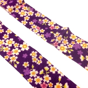 May include: Two purple fabric ribbons with a white floral pattern. The ribbon has a textured surface and a repeating pattern of white flowers with yellow centers and pink accents.