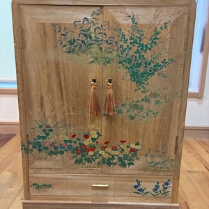 May include: Wooden cabinet with floral artwork in shades of green, red, yellow, and blue. The cabinet has two doors with gold tassel pulls and a drawer with a gold handle. The cabinet is made of light-colored wood.