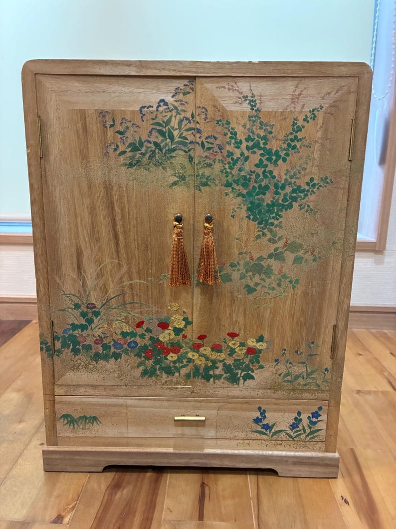 May include: Wooden cabinet with floral artwork in shades of green, red, yellow, and blue. The cabinet has two doors with gold tassel pulls and a drawer with a gold handle. The cabinet is made of light-colored wood.