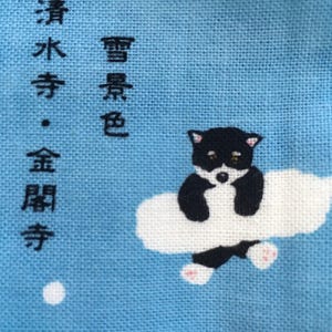 Op de afbeelding: Een blauwe stof met witte wolken en een zwart-witte pandabeer die op een wolk zit. De stof heeft Japanse tekst die "Winter, helder water, gouden haan" luidt.