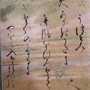 May include: Close-up of a traditional Japanese artwork featuring calligraphy in black ink on a beige and green background. The artwork has a textured appearance and is framed with a patterned border.