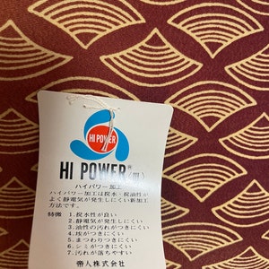 May include: A white tag with the text "HI POWER" in blue and red lettering. The tag is attached to a red fabric with a pattern of yellow and brown overlapping semi-circles. The tag describes the "HI POWER" processing, which is a water-repellent, oil-resistant, and anti-static treatment. The tag also lists the benefits of the processing, including water resistance, static resistance, oil resistance, dust resistance, and stain resistance. The tag is made by Teijin Limited.