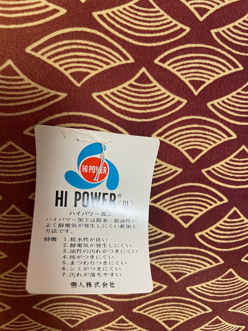 May include: A white tag with the text "HI POWER" in blue and red lettering. The tag is attached to a red fabric with a pattern of yellow and brown overlapping semi-circles. The tag describes the "HI POWER" processing, which is a water-repellent, oil-resistant, and anti-static treatment. The tag also lists the benefits of the processing, including water resistance, static resistance, oil resistance, dust resistance, and stain resistance. The tag is made by Teijin Limited.