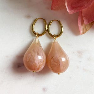 Op de afbeelding: Een paar gouden oorringen met peervormige, perzikkleurige hars hangers. De hangers hebben een gemarmerd effect. De oorbellen worden getoond op een wit marmeren oppervlak. De hangers zijn ongeveer 3 cm lang.