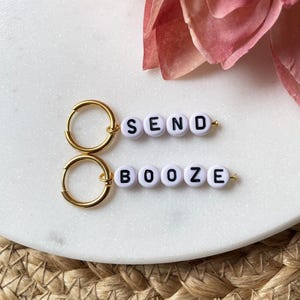 Op de afbeelding: Twee goudkleurige oorringen met witte letterkralen die "SEND" en "BOOZE" spellen. De oorbellen worden gepresenteerd op een wit marmeren oppervlak, met een roze bloem op de achtergrond.