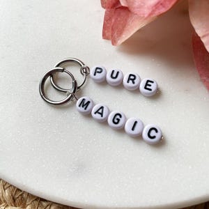 May include: Silver hoop earrings with white letter beads spelling "PURE MAGIC". The earrings are displayed on a white marble surface, with a pink flower in the background. A simple and stylish accessory.